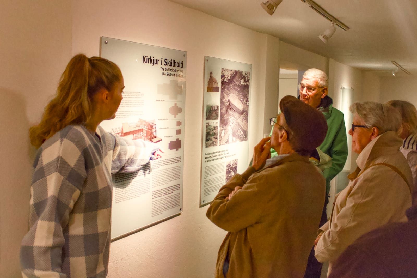 A guide explaining Skálholt archaeology panels to visitors inside the historic site exhibition area.