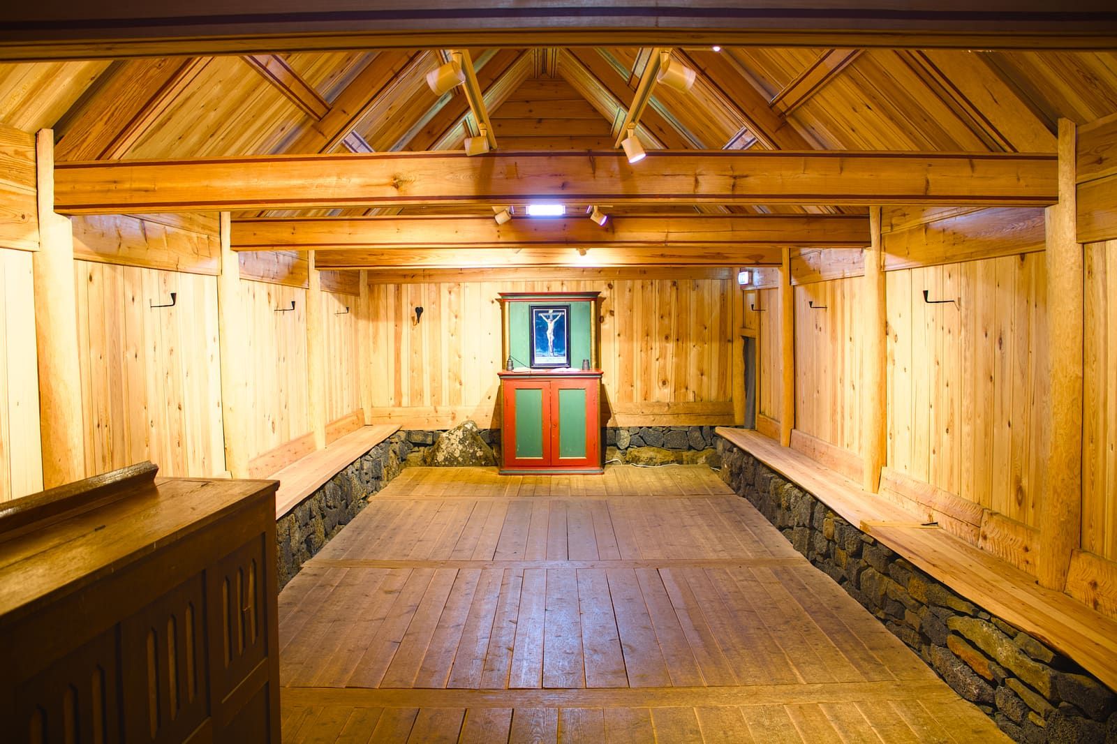 Wooden interior of Þorláksbúð at Skálholt with benches, timber walls, stone base, and a small altar.