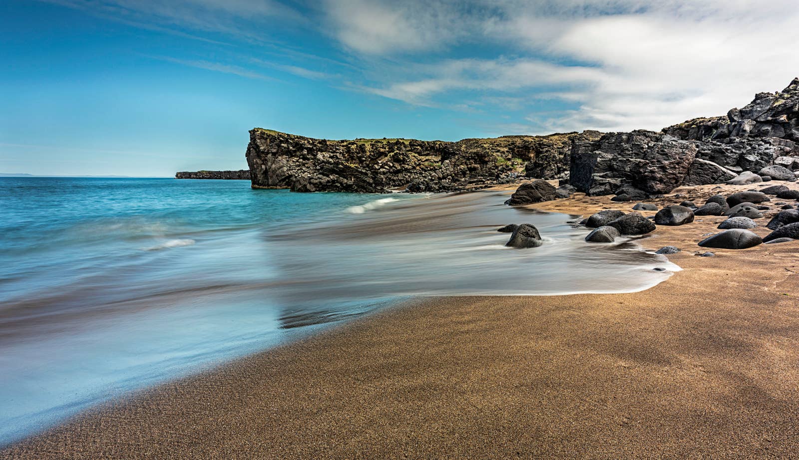 Golden sand and blue water inside Skarðsvík cove below black lava rock.
