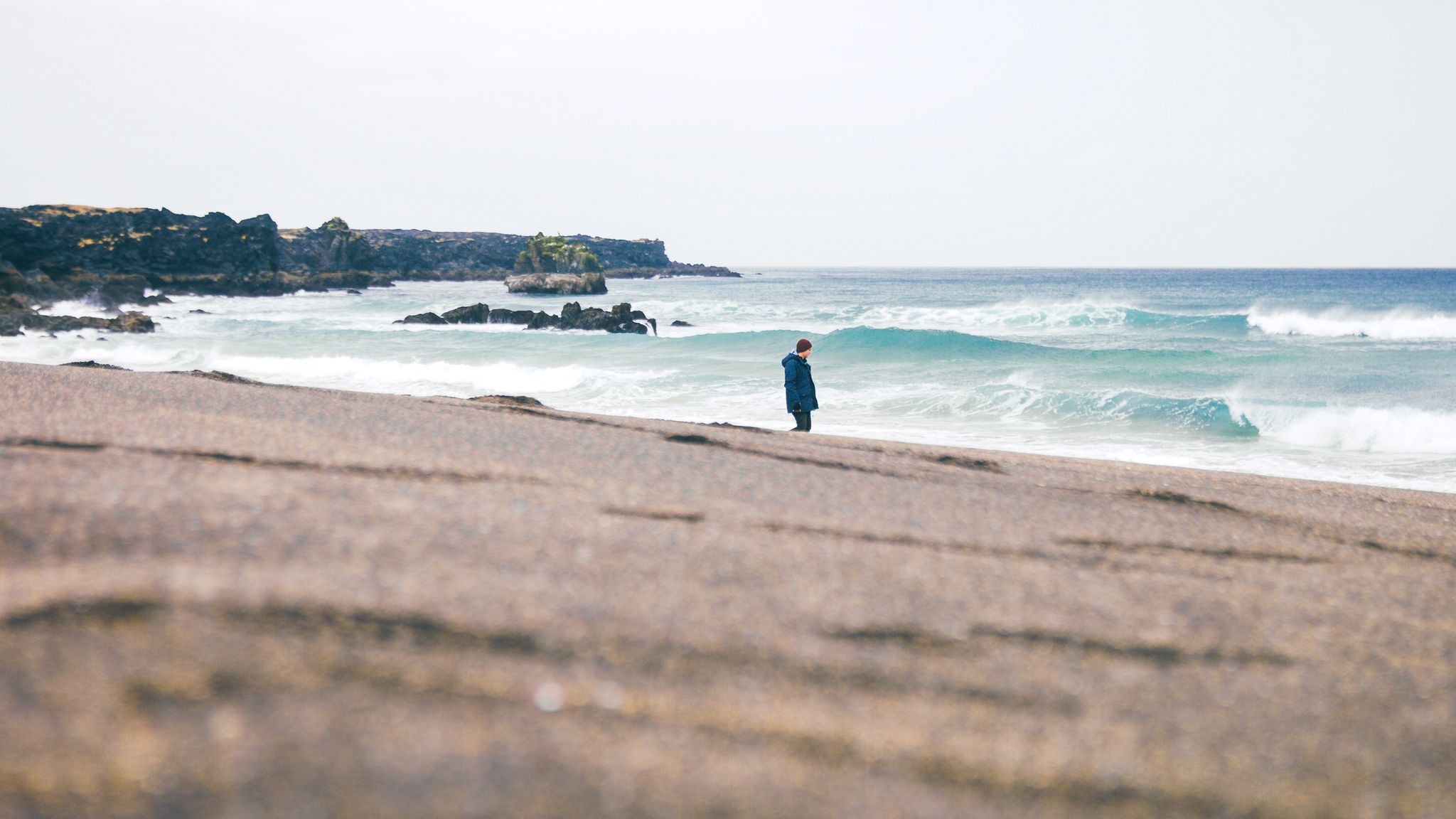 Visitor standing on Skarðsvík Beach with yellow sand, turquoise surf, and dark lava cliffs.