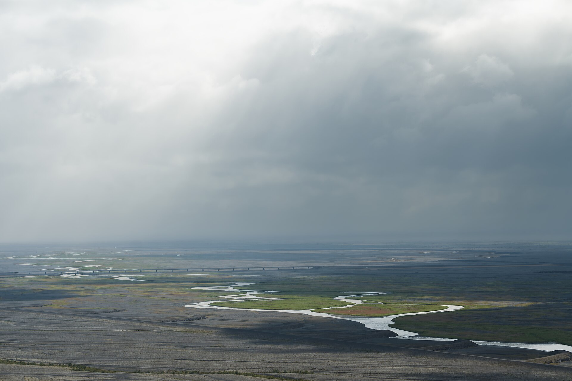Distant bridge and braided rivers across the dark plain of Skeiðarársandur under low clouds.