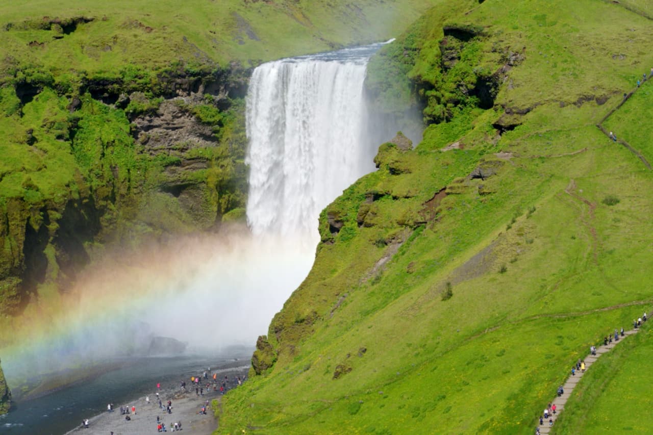 Rainbow mist at Skogafoss with the stair route rising beside the waterfall.