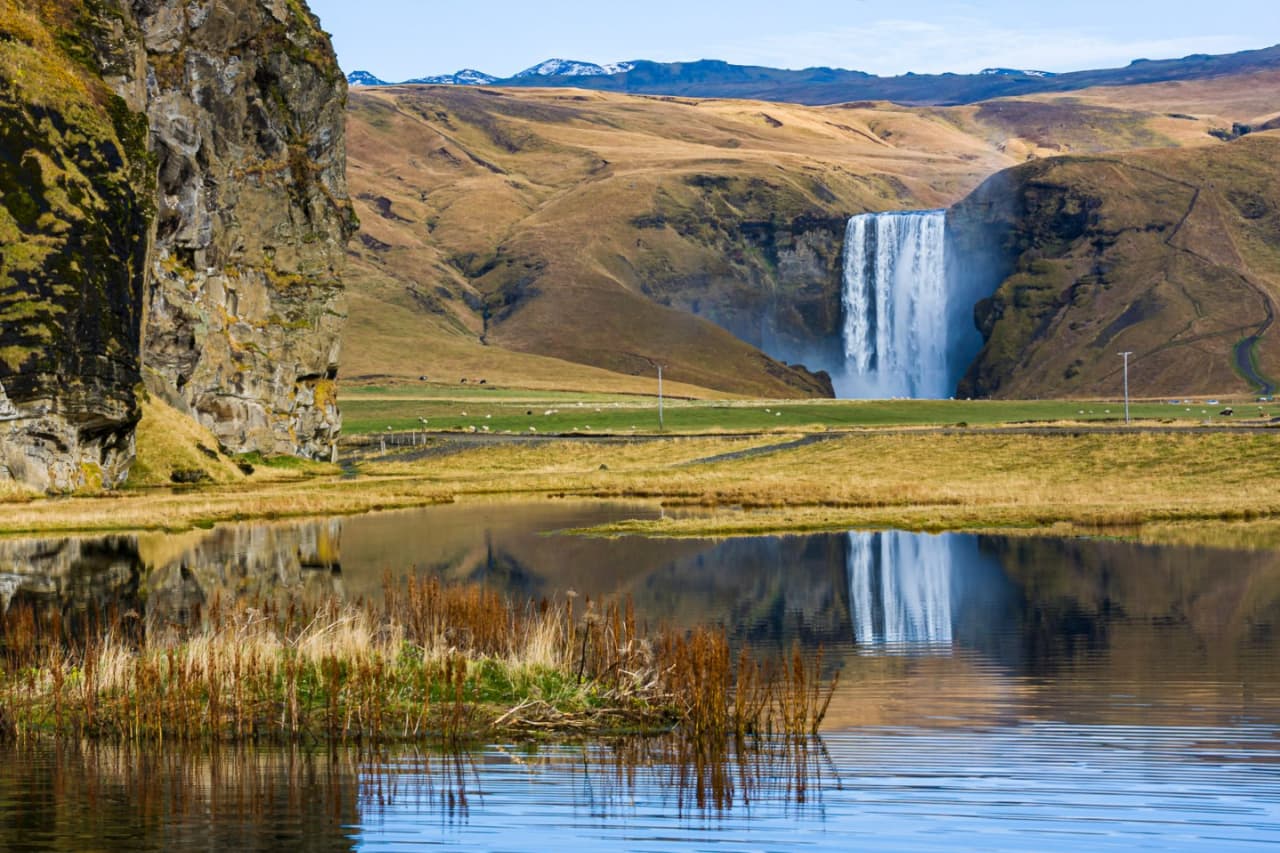 Skogafoss reflected in wet ground below green cliffs in South Iceland.