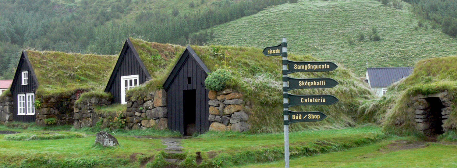 Turf-roofed buildings and visitor signs at Skógar Museum below green South Coast slopes.