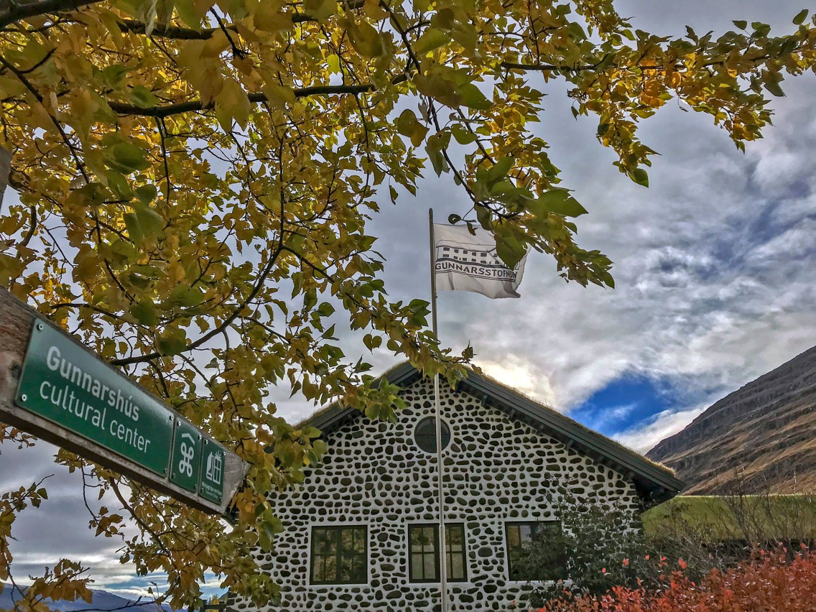 Gunnarshús cultural center sign, Skriðuklaustur flag, and the turf-roofed house framed by autumn leaves.