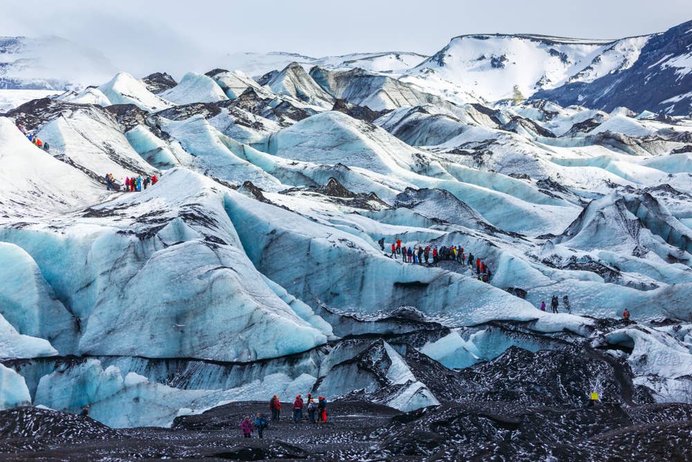 Guided group walking across ash-striped Sólheimajökull Glacier with mountain walls behind the ice
