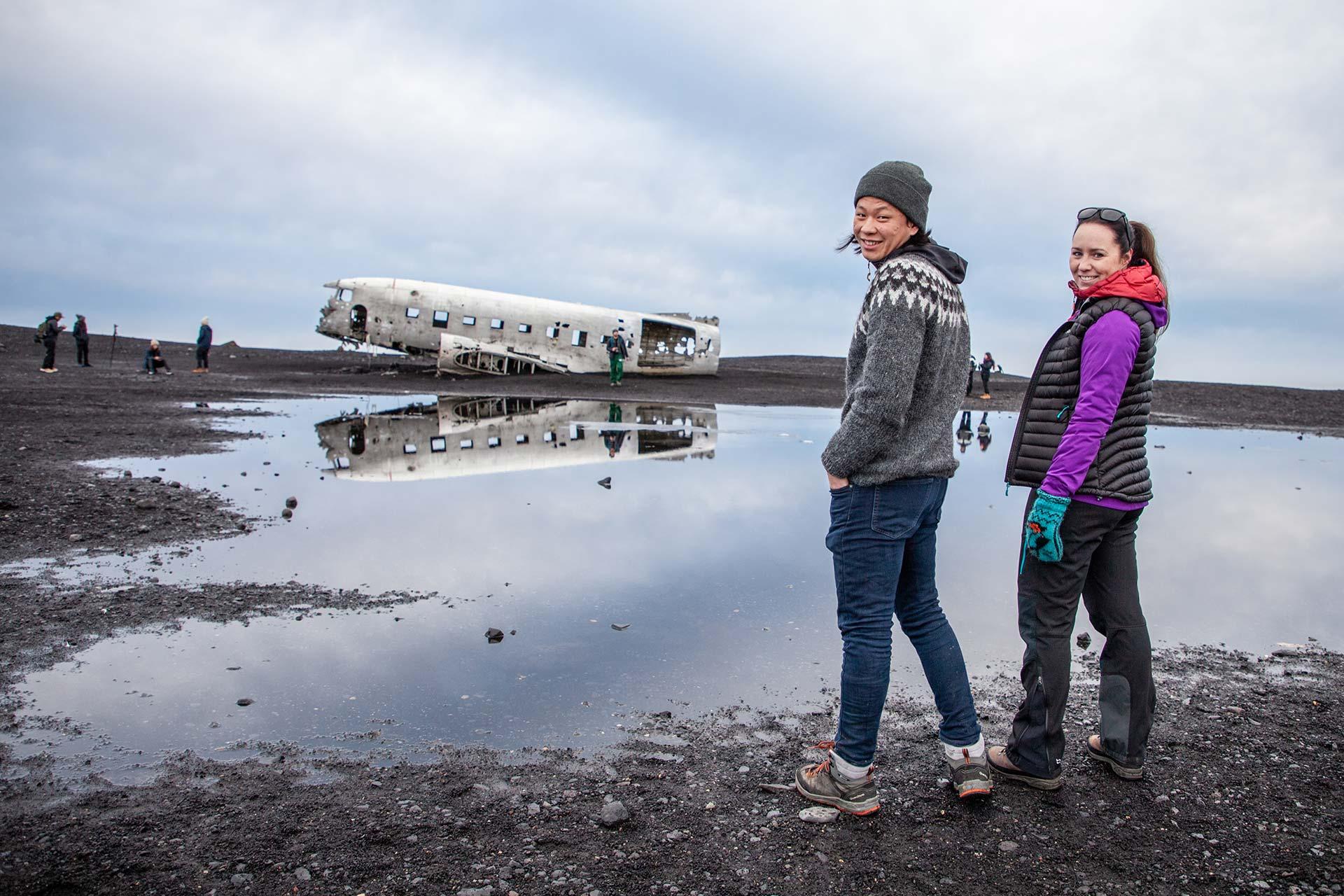 Visitors standing near shallow water with the Sólheimasandur plane wreck behind them.