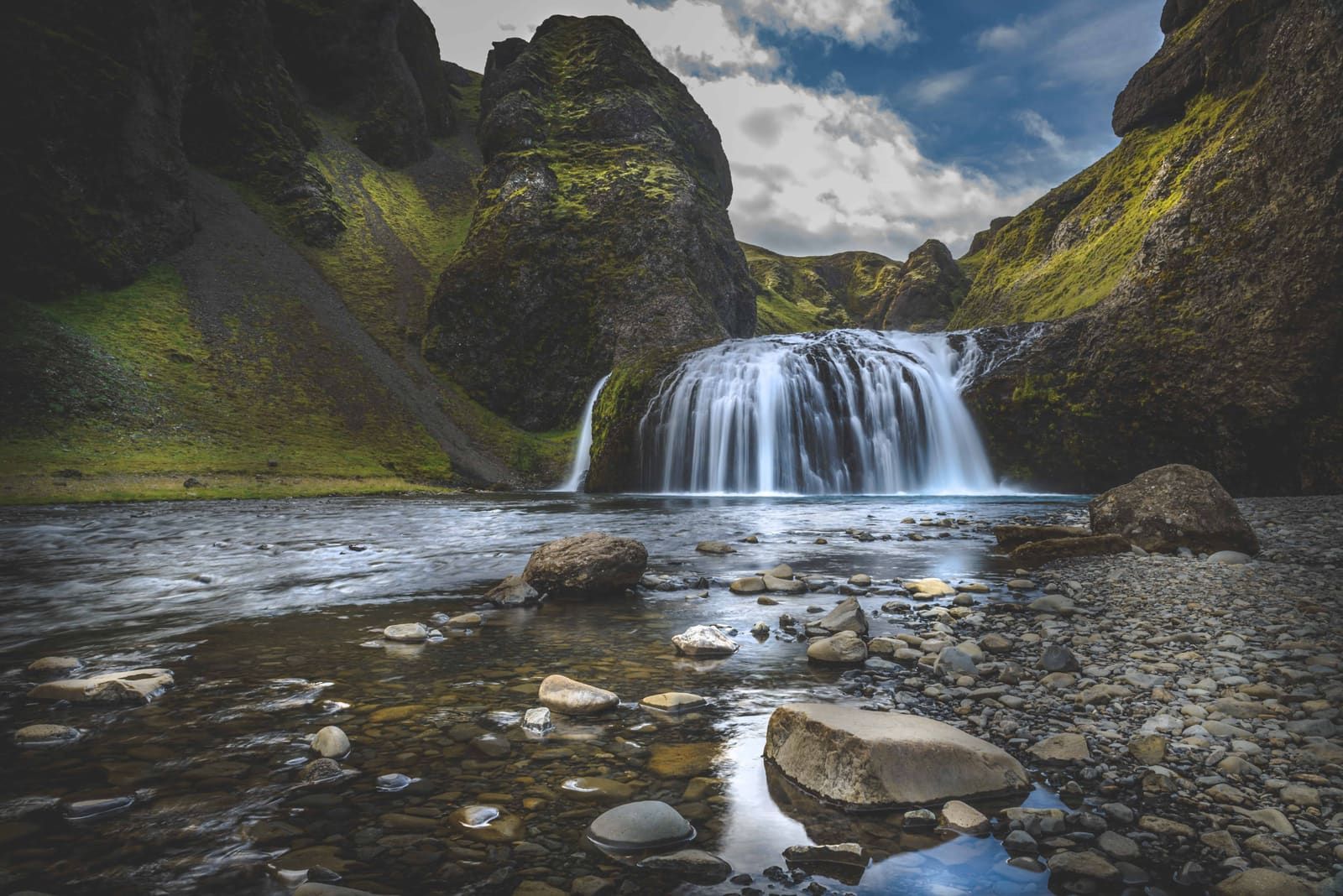 Wide river-level view of Stjórnarfoss with the lower curtain drop and mossy cliffs rising behind it.