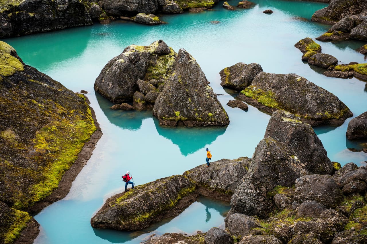 Hikers crossing giant mossy boulders beside bright blue-green ponds in Stórurð below Dyrfjöll in East Iceland.