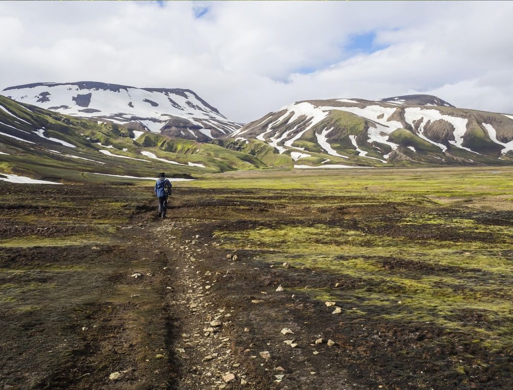 A lone hiker crossing dark highland ground toward snow-patched hills near Strútslaug.