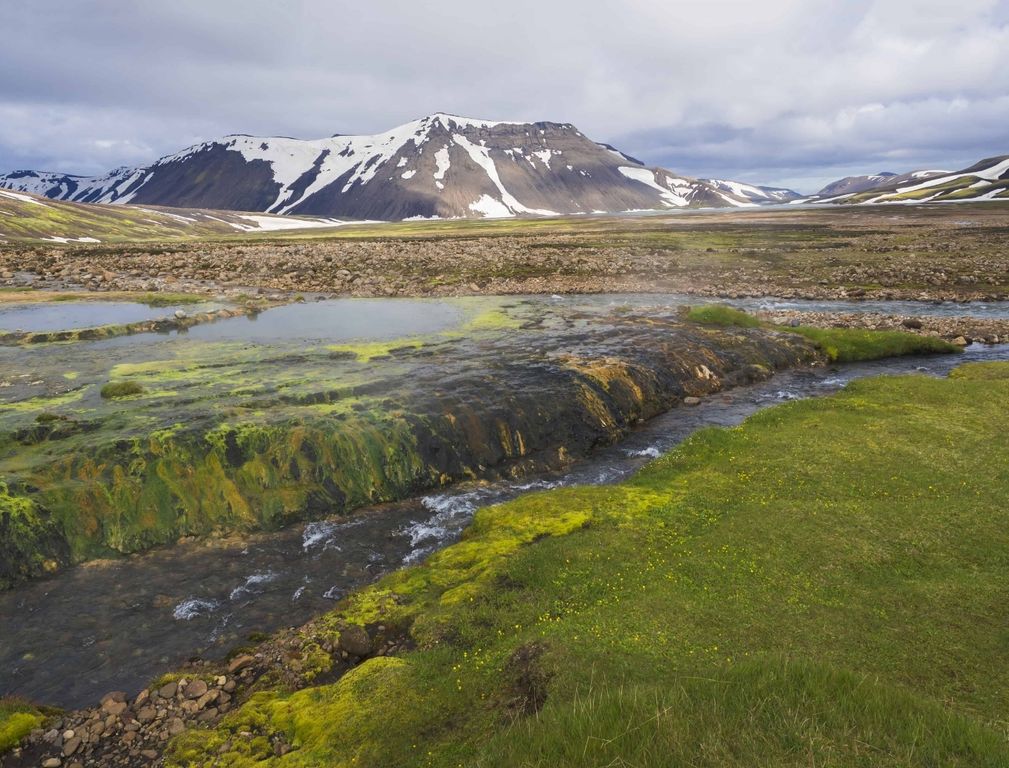 The Strútslaug pool edge beside a narrow river with snow-streaked highland mountains beyond.