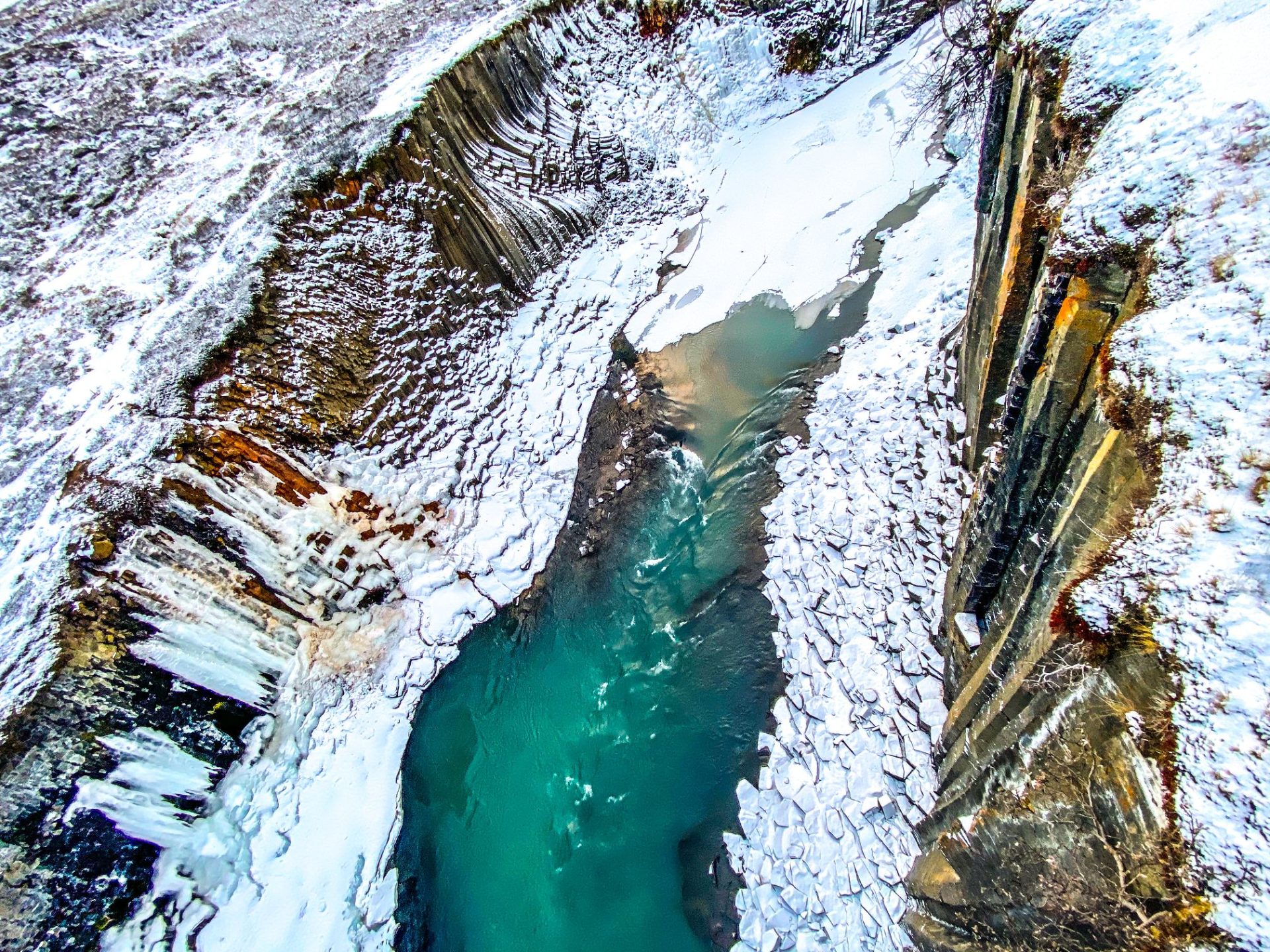 Top-down view of the blue river threading between basalt walls at Stuðlagil Canyon in snowy conditions.