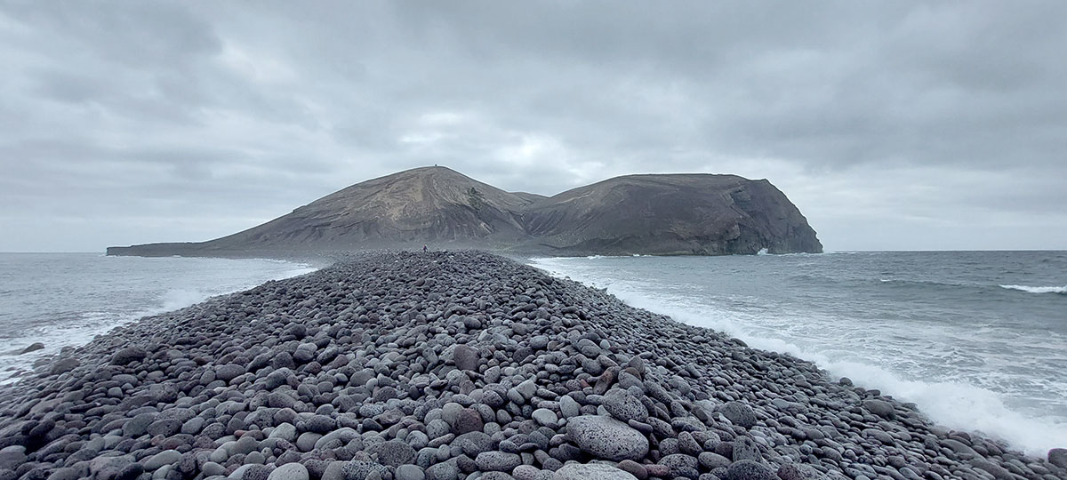 Surtsey seen across a black cobble spit with waves on both sides under grey clouds.