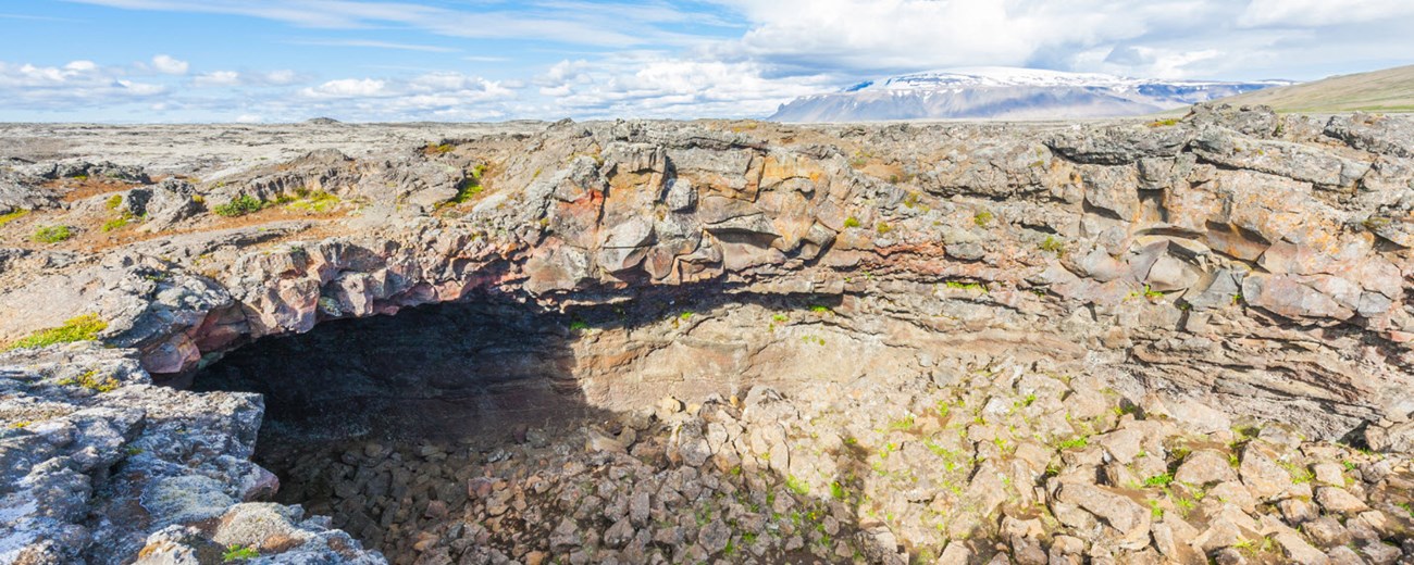 Surtshellir cave opening in a rough lava field with pale sky and distant highland slopes.