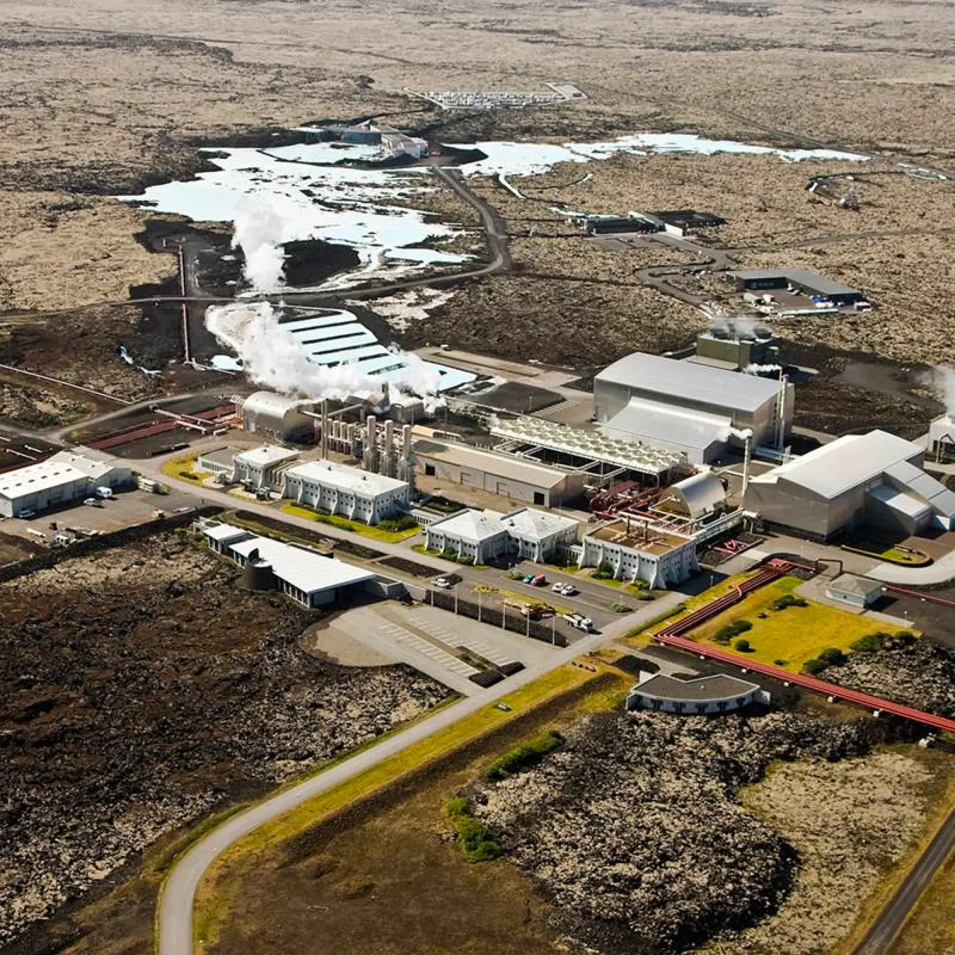 Aerial view of Svartsengi Power Station with steam, roads, buildings, black lava, and pale geothermal water.