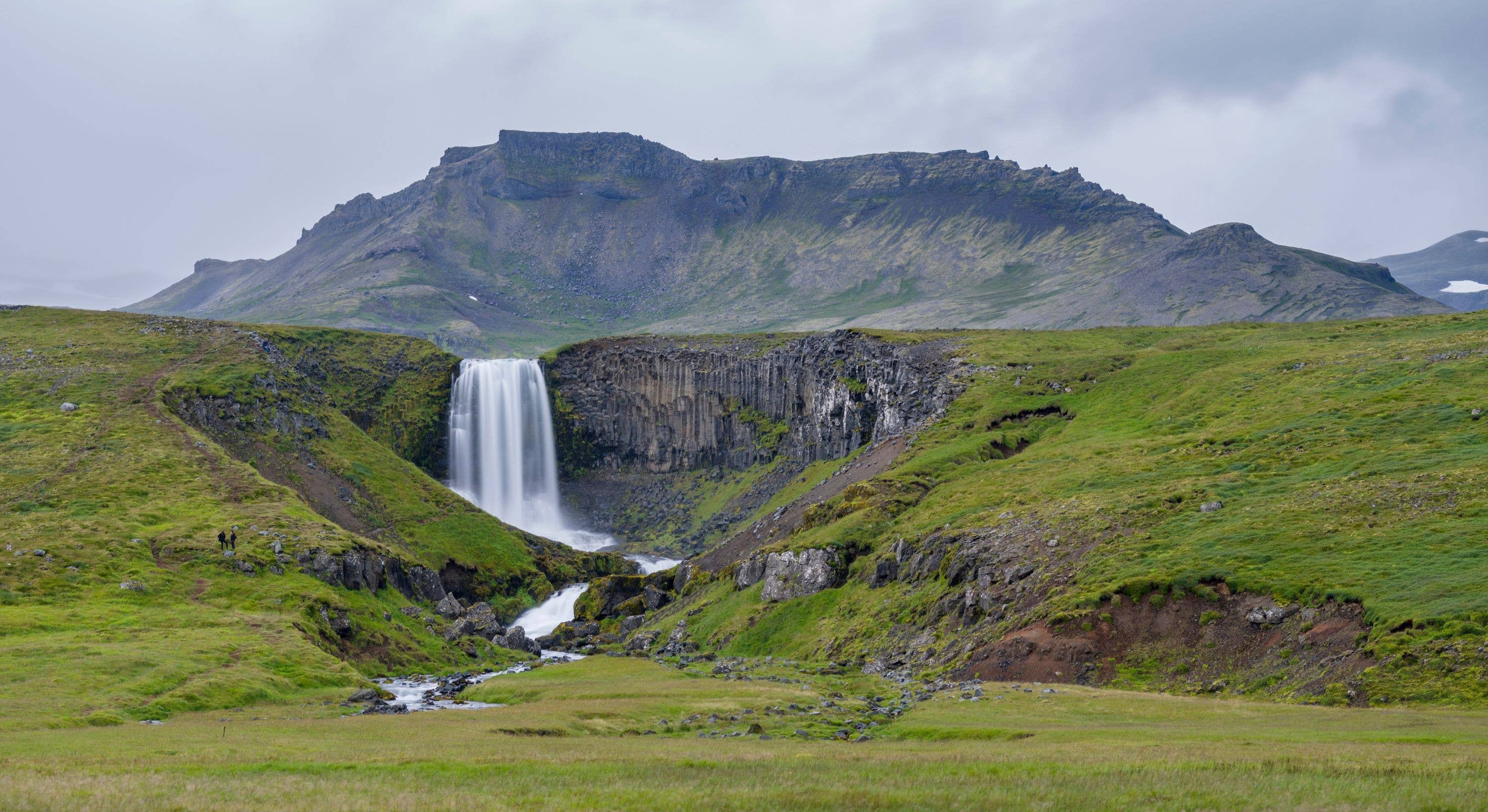 Svöðufoss Waterfall dropping over basalt columns with green slopes and a mountain ridge behind it on Snæfellsnes.