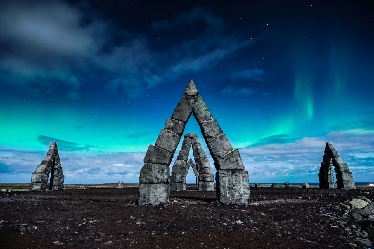 Stone arches of the Arctic Henge under a blue night sky with northern lights above Raufarhöfn.