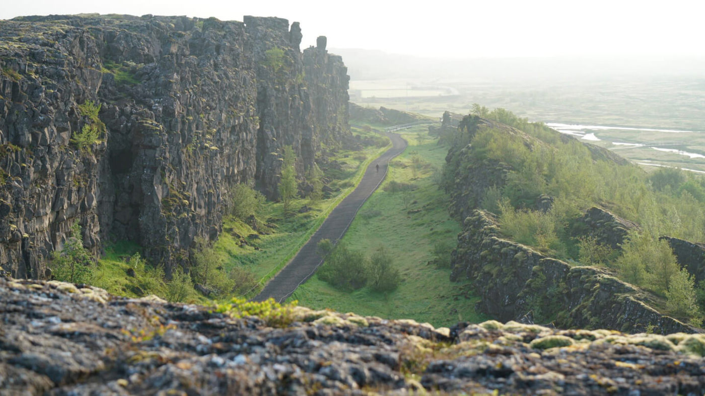 Marked path through dark rift cliffs at Þingvellir with open lowland and water beyond.