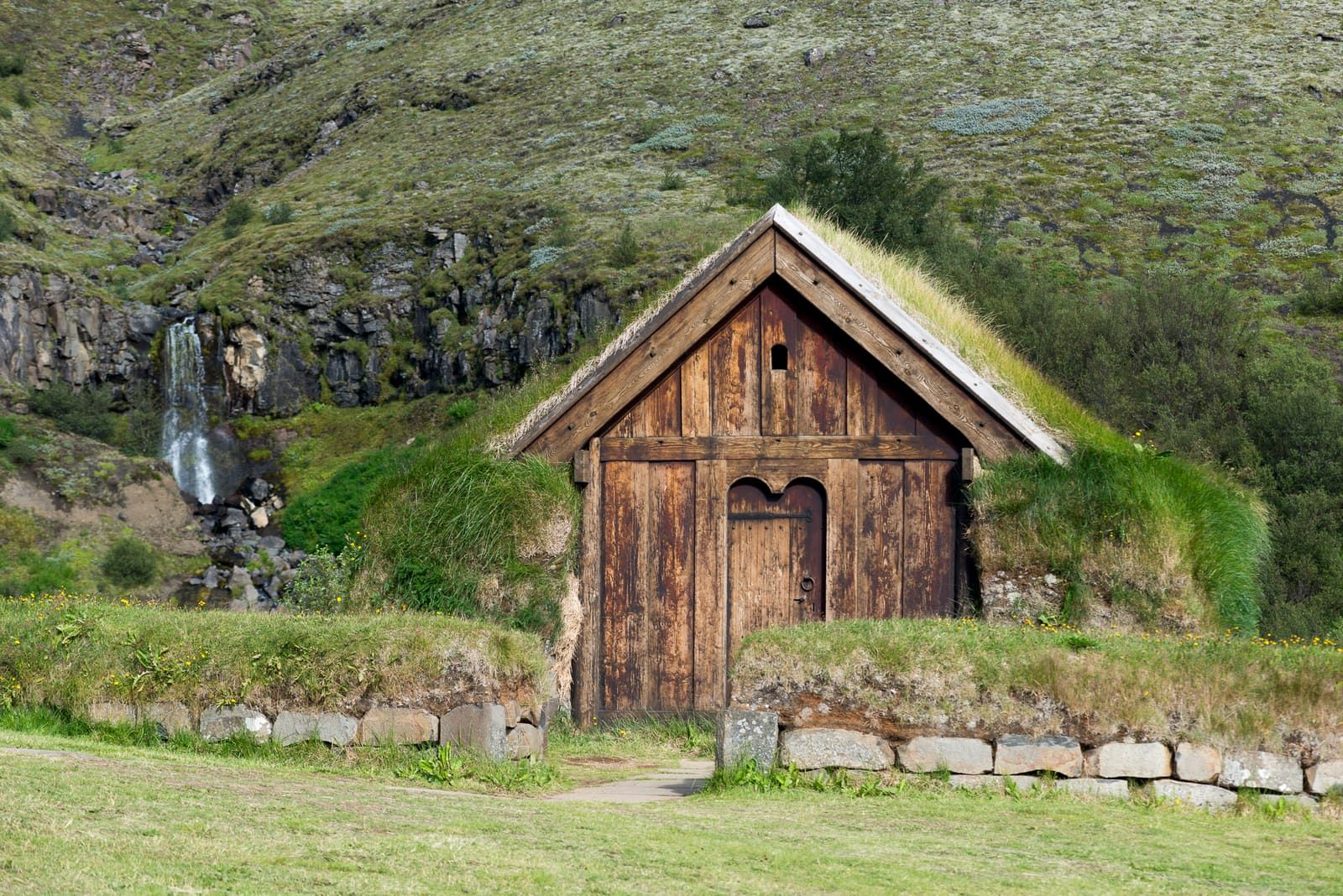Small turf-roofed church beside Þjóðveldisbærinn with a hillside and waterfall behind it.