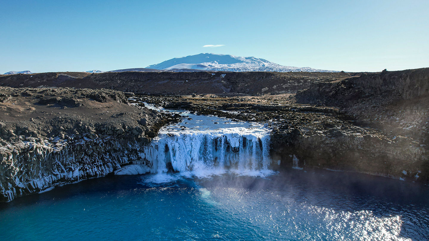 Þjófafoss waterfall on Þjórsá with snowy Hekla mountain visible beyond dark lava fields.