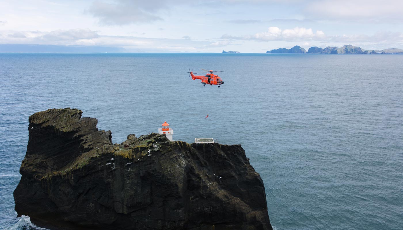 Helicopter hovering above Þrídrangaviti on its isolated sea stack with the Westman Islands on the horizon.