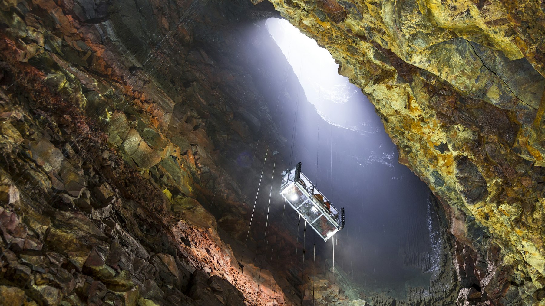Cable lift descending through the opening of Þríhnúkagígur into a broad mineral-colored magma chamber.
