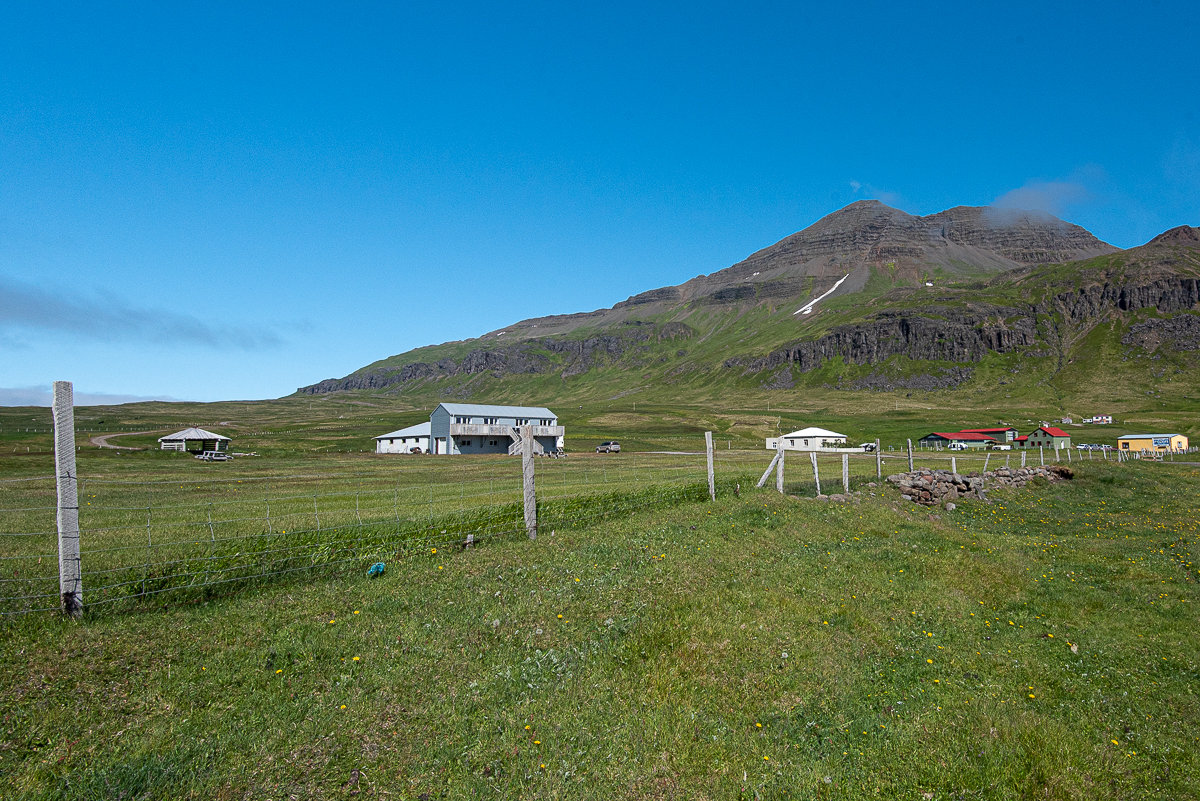 Small buildings and fenced grassland below a steep mountain in the Trékyllisvík area of Strandir.