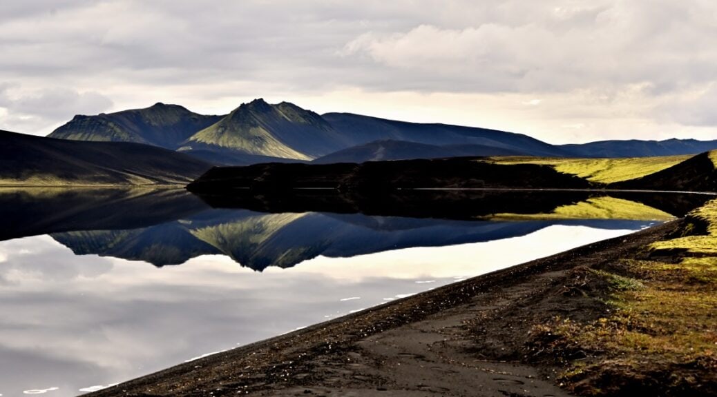 Calm Grænavatn water in the Veiðivötn area reflecting dark highland mountains under a cloudy sky.