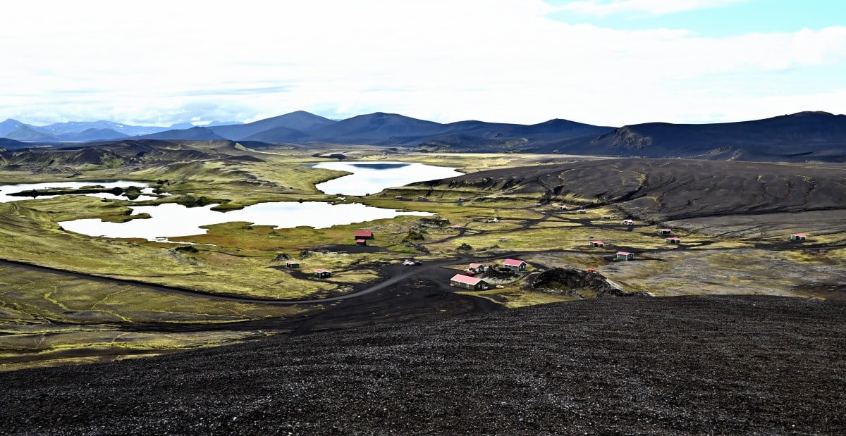 View over Veiðivötn lakes, black volcanic slopes, small red-roofed huts, tracks, and distant highland mountains.