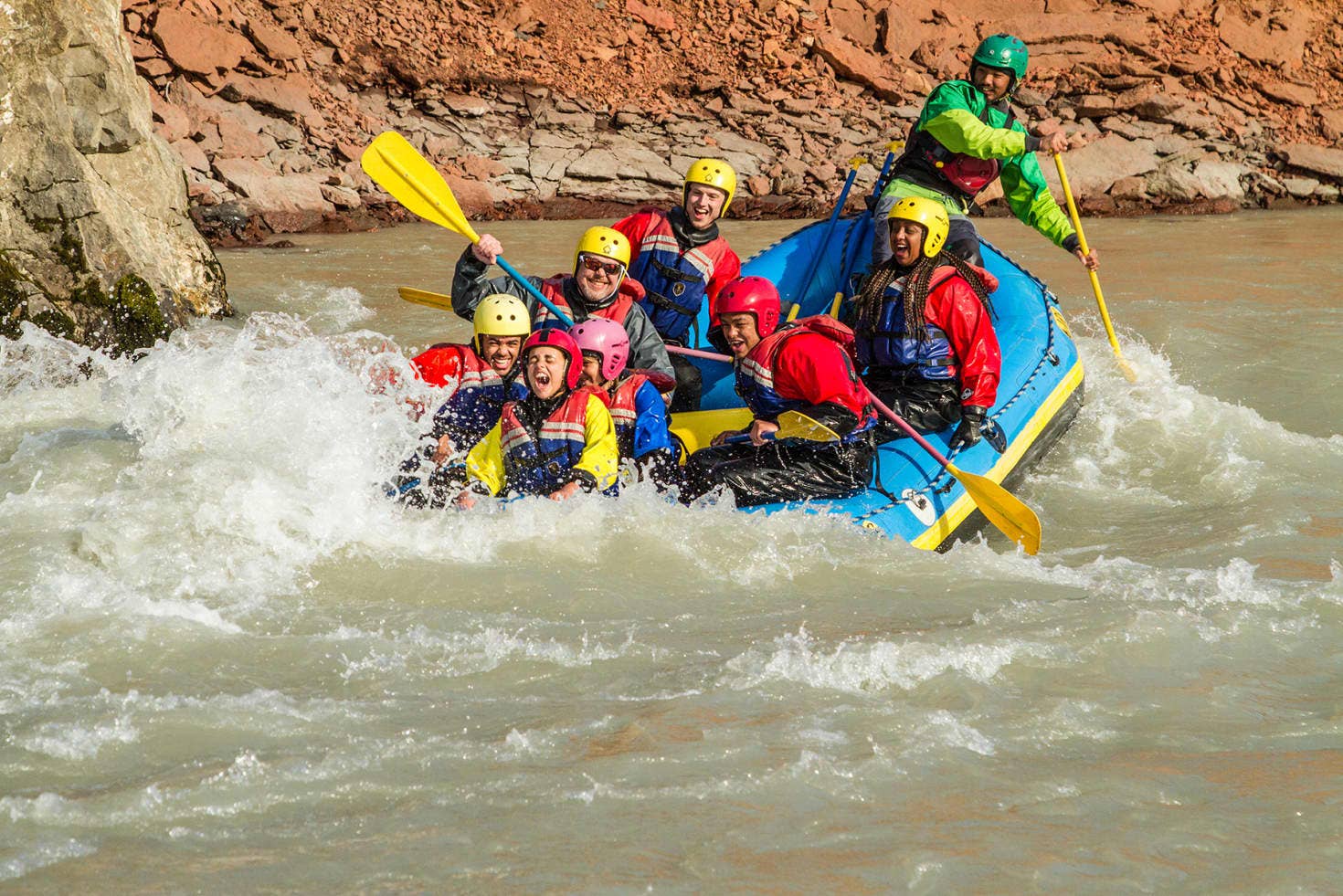 A guided raft with families paddling through pale Vestari Jökulsá water beside reddish canyon rock.
