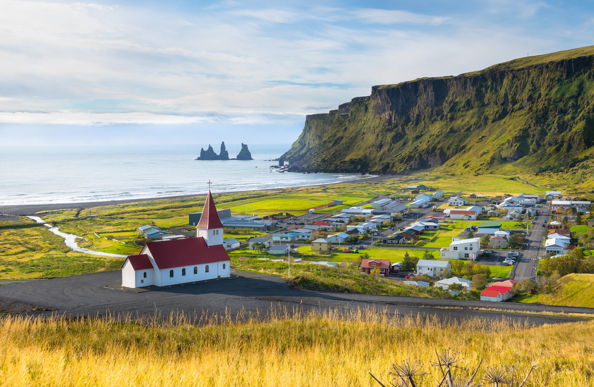 Vík village with Víkurkirkja, black-sand coast, and Reynisdrangar in the distance.