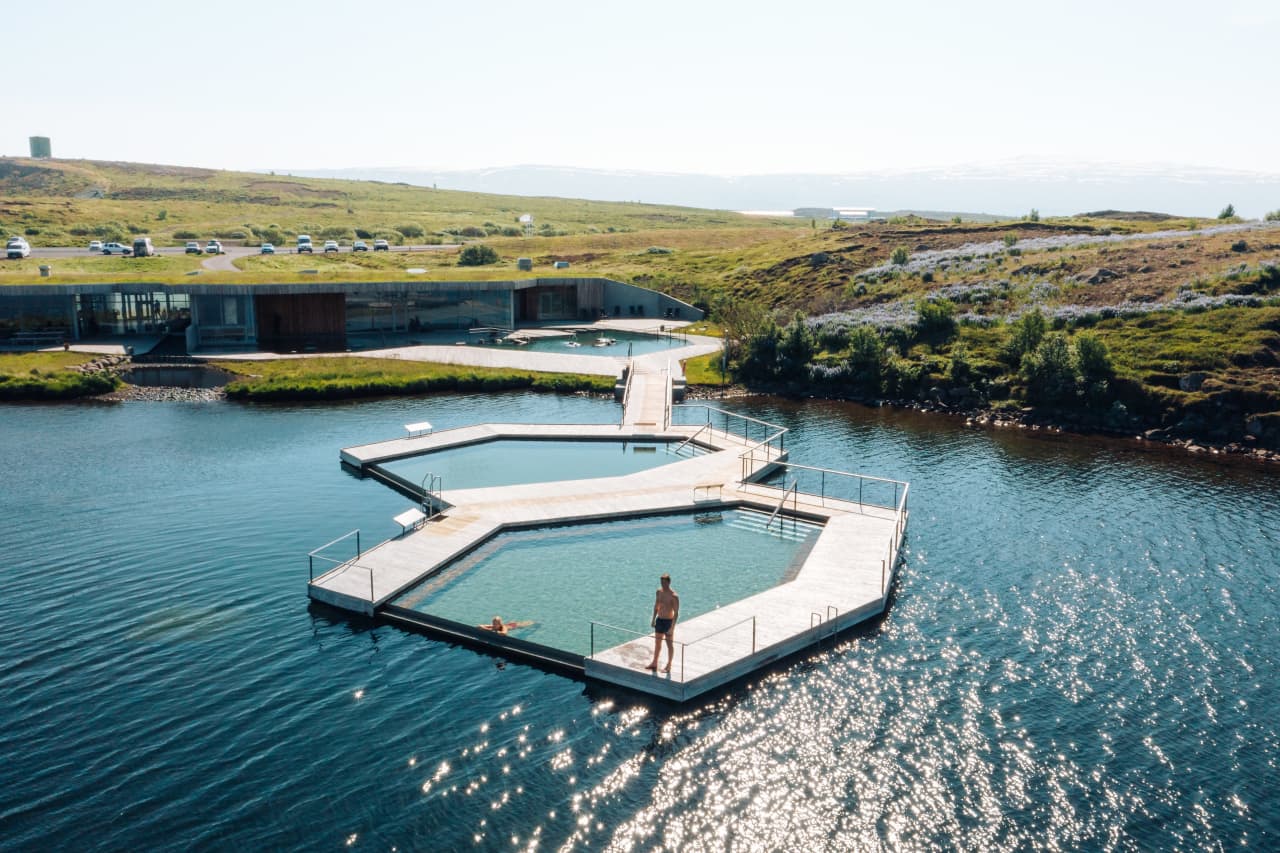 Wide summer view of the floating pools at Vök Baths with the low main building on the shore behind them.