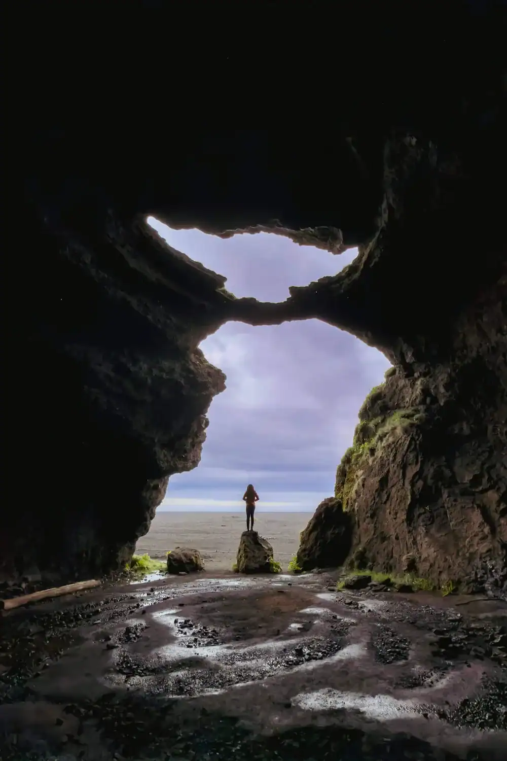 Person standing inside Gígjagjá under the Yoda-shaped cave opening at Hjörleifshöfði.